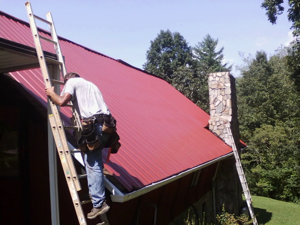 Roofer on a ladder beside a red metal roof and stone chimney — Soffit & Fascia Repair by Roofing Brothers in Rockaway
